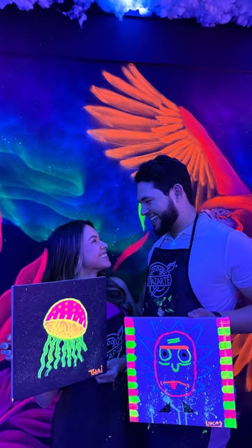 Casal sorrindo com duas telas pintadas sob iluminação neon